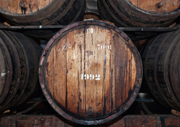 A large wooden barrel labeled "1992," "700L," and "610L," sits on a rack among other barrels in a dim storage setting. Stains cover the barrel's surface, indicating age and use.